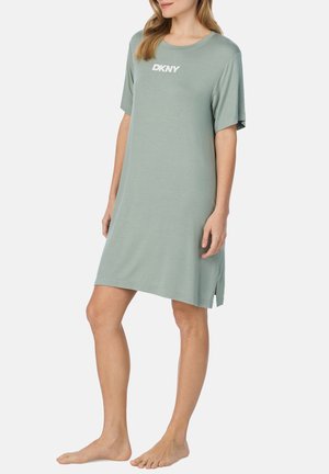 Kobieta w luźnej, krótkoszlfowej sukience w kolorze szałwii z logo "DKNY" na piersi, stojąca boso na tle jednolitej białej ściany.