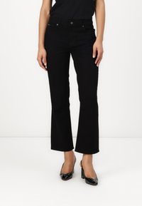 Sieviete, tērpta melnās bootcut džinsās un melnos lakādos augstpapēžu sandalos, stāvot pret baltu fona.