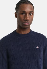 Marineblauer Kabelstrickpullover aus Wolle mit Rundhalsausschnitt und kleinem gestickten Logo auf der Brust.