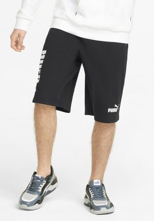 Męskie nogi w czarnych szortach Puma i szarych butach sportowych Puma, stojące na białym tle.