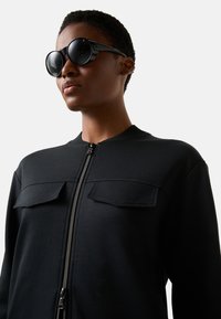 Schwarzer Sweatshirt mit Reißverschluss und zwei Fronttaschen, aus glattem Stoff. Kombiniert mit übergroßen schwarzen Sonnenbrillen in runder Form.