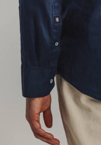 Donkerblauw linnen overhemd met een knoopsluiting bij de manchet, met een klein merkje. Glad textuur, getailleerde pasvorm, zichtbare knoopdetails.