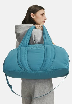 Žena v šedé kapuci drží velkou modrou polstrovanou sportovní tašku přes rameno na bílém pozadí.