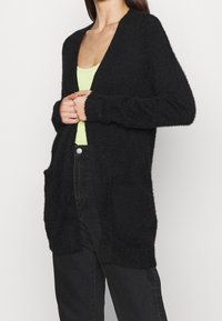 Svart, fluffig cardigan med långa ärmar, öppen framsida och två framfickor. Tillverkad av mjukt, texturerat material, designad för en avslappnad passform.