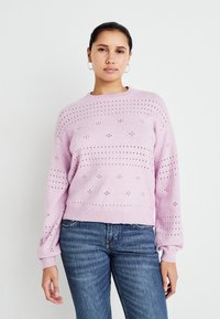 Pulover tricotat de culoare roz deschis, cu guler rotund, având modele cu ochiuri și manșete cu nervuri, asortat cu blugi albaștri.