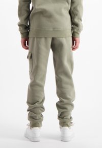 Persoon in olijfgroene trainingsbroek en bijpassend sweatshirt, staande met ontspannen handen, draagt witte Nike Air sneakers.