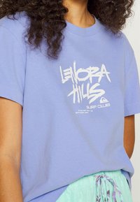 Camiseta de algodón de color púrpura claro con texto gráfico en blanco "LENORA HILLS SURF CLUB" y texto adicional debajo. Manga corta y escote redondo.