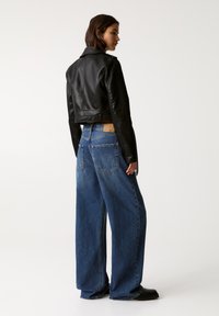 Giacca nera in pelle a maniche corte con colletto a revers, abbinata a jeans in denim blu con gamba larga, caratterizzati da cuciture visibili e tasche posteriori.