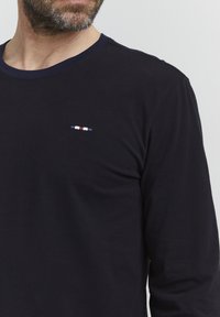 Tengerészkék hosszú ujjú póló, pamutból készült, kerek nyakkivágással és egy kis hímzett logóval, amely piros, fehér és kék részletekkel díszíti a mellrészt.