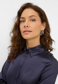 Navy Satinhemd mit spitzem Kragen und Knopfleiste. Das Modell trägt einen großen, dekorativen Ohrring mit floralem Design.