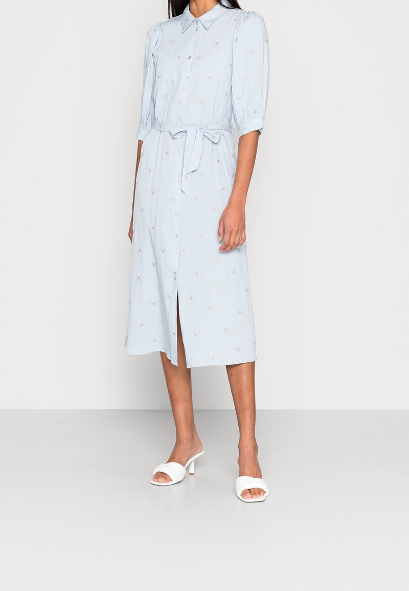 Femme portant une robe midi bleu clair à boutons, ceinture à la taille et manches bouffantes trois-quarts, associée à des sandales blanches à talons et à bout ouvert.