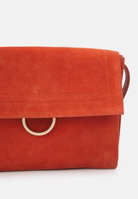 Rode suède handtas met een klep en een ronde gouden ringaccent, met zichtbare stiksels langs de randen. Compacte rechthoekige vorm.