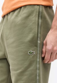 Κοντινό πλάνο ενός ατόμου που φοράει ελαιχρώς πράσινα παντελόνια φόρμας Lacoste με το λογότυπο κροκόδειλου και ραμμένη ρίγα στην πλευρά, το χέρι ακουμπάει πάνω στον μηρό.