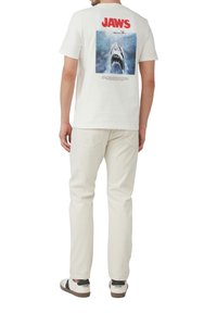 Biała koszulka z krótkim rękawem z grafiką na plecach przedstawiającą rekina. Napis "JAWS" na czerwono, wyrażony dużą czcionką. Uzupełniona jasnymi spodniami i codziennymi butami.