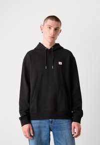 Zwarte hoodie van katoen, met een voorzak, trekkoorden en een klein logopatch op de borst. Draagt over een blauwe jeans.