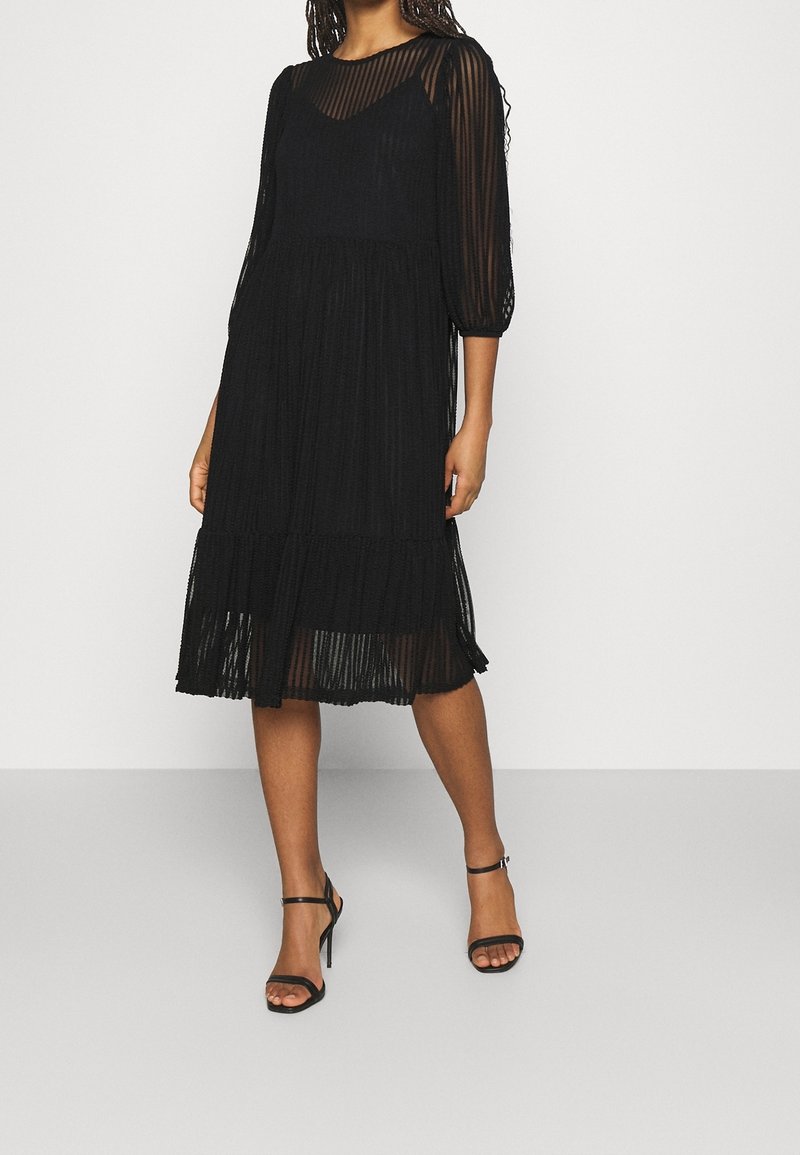 Femme portant une robe noire plissée longueur genou avec des manches trois-quarts transparentes et des sandales à talons hauts noires à lanières sur un fond clair.