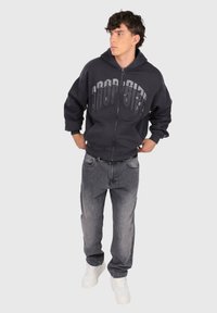 Ung mand står med hænderne i lommerne, iført en sort zip-up hoodie med teksten "DROP GIFT", grå jeans og hvide sneakers.