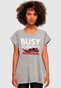 Šedé oversized tričko s bílým a červeným grafickým nápisem "BUSY LOOKING DEADCOOL" s karikaturami. Měkká textura, krátké rukávy.