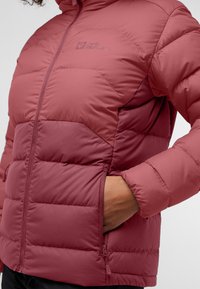 Veste doudoune rouge avec des sections matelassées et une poche zippée. Le matériau semble lisse avec un fini mat et un logo sur la poitrine.