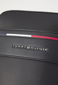 Musta rakenteinen materiaali, jossa on erottuva punainen, valkoinen ja sininen raita. Sisältää "TOMMY HILFIGER" -logon hopeana kohotettuna tasaiselle paneelille.
