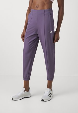 Osoba nosząca fioletowe spodnie dresowe Adidas oraz białe buty sportowe Adidas stoi na tle jednolitego tła.