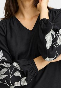 Blouse noire avec un décolleté en V, en tissu texturé et manches longues bouffantes ornées de broderies blanches en forme de feuilles.