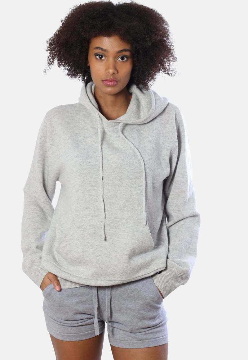 Giovane donna con capelli ricci che indossa un felpa leggera grigia e pantaloni corti abbinati, in piedi con le mani nelle tasche su uno sfondo bianco.