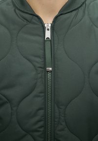 Gros plan sur la fermeture éclair d'une veste matelassée vert foncé, montrant des motifs de couture courbés et une languette en tissu sur le curseur métallique de la fermeture.