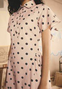 Vestido de manga corta con cuello en color rosa claro y grandes lunares negros. Cuenta con cierre de botones en la parte delantera y paneles laterales recogidos.