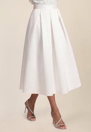 Weißer plissierter Midirock kombiniert mit silbernen, römisch geschnürten High-Heel-Sandalen, getragen von einer Person vor einem einfarbigen Hintergrund.