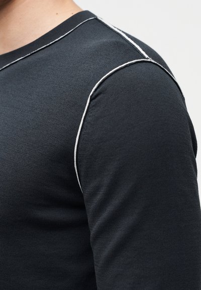 Gros plan d'une personne portant une chemise à manches longues foncée avec des coutures contrastantes blanches le long de l'épaule et de l'encolure.