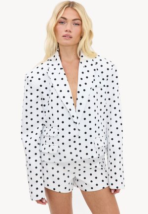 Femme aux cheveux blonds portant un ensemble blazer blanc et short à pois noirs, debout devant un fond uni.