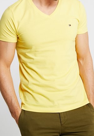 Man wearing a fitted yellow V-neck T-shirt with a small logo on the chest and olive green pants with hands in pockets.