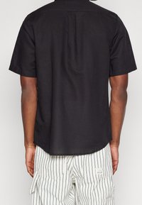 Chemise noire à manches courtes en tissu léger, avec un col classique, un pli dans le dos et un ourlet arrondi.