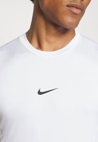 Kratek rokav, bel športni majica iz zračnega materiala, ki ima majhen črn Nike logotip na sredini prsnega dela. Gladka tekstura.