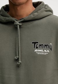 Sudadera verde con cordón, que presenta un destacado logo blanco en el pecho que dice "Tommy Jeans, N.V." Tejido suave, corte relajado.