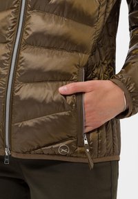 Vadderad jacka i olivgrön, med en glänsande yta, silverfärgade dragkedjor och en sidoficka. Anmärkningsvärd logotypdetalj nära fållen.