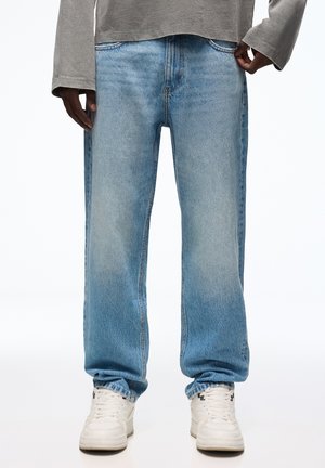 Person iført lyseblå løstsiddende jeans og hvide sneakers, stående med hænderne tæt på lommerne mod en ensfarvet baggrund.