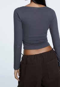 Vrouw in een strakke, lange mouwen grijze crop top en losse donkerbruine broek met grote achterzakken, van achteren getoond tegen een effen achtergrond.