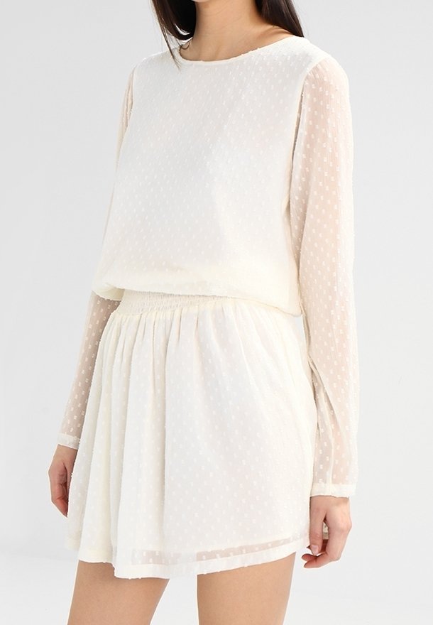 Femme portant un chemisier blanc texturé à manches longues et une jupe assortie avec une taille cintrée sur un fond uni.