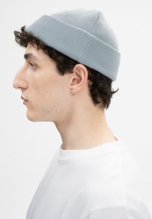 Junger Mann mit lockigem Haar, der eine hellgraue Mütze, kleinen Creolenohrring und ein weißes Rundhalsshirt trägt, schaut nach links vor einem weißen Hintergrund.