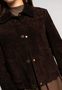 Chaqueta de ante marrón con cuello, que cuenta con dos bolsillos frontales y botones a presión plateados. Superficie texturizada con un acabado suave y mate.