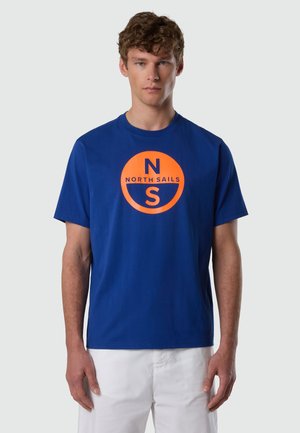 Jeune homme portant un t-shirt bleu avec un logo North Sails orange et un pantalon blanc, debout devant un fond gris clair uni.