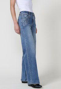 Pantalones vaqueros de mezclilla azul, acampanados, con detalles desgastados, talle alto y diseño de pierna recta. Cuenta con bolsillos y costuras en contraste.