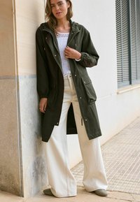 Trench coat verde oliva con cappuccio, maniche lunghe, due tasche frontali e una cerniera. Abbinato a pantaloni crema a gamba larga e scarpe grigie morbide.