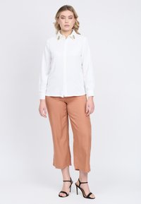 Camicia bianca con pulsanti e decorazioni dorate sul colletto, abbinata a pantaloni a gamba larga color ruggine e sandali neri con tacco.