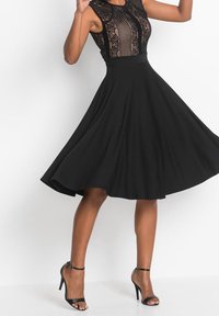 Černé šaty s přiléhavým krajkovým korzetem, rozšířenou sukní a hladkým materiálem; doplněné černými sandály na tenkém podpatku.