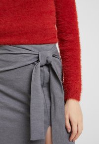 Un pull rouge duveteux associé à une jupe portefeuille grise avec un détail de nouage et une fente sur le côté. Textures douces et tissu lisse visibles.