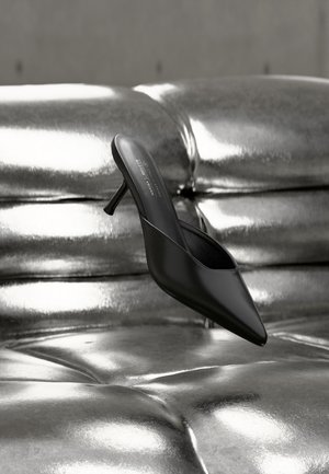 Zwarte mule met puntige neus en een smalle hak, geplaatst op een glanzend, metallic zilveren gekussende ondergrond.