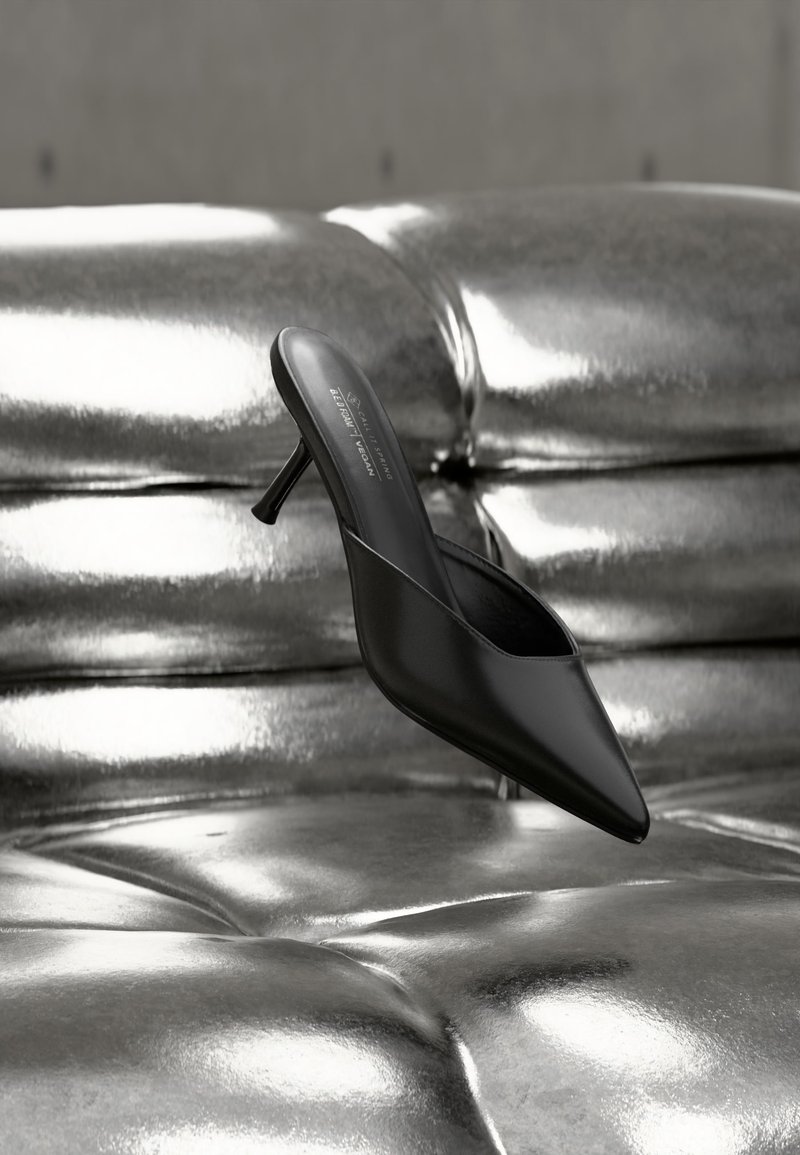 Mule noire à bout pointu avec un talon fin, posée sur une surface matelassée argentée brillante et métallique.